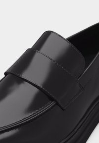 Mocassin en cuir noir poli avec une large lanière sur le dessus et des coutures visibles le long des bords.