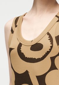 Εγγύς λήψη ατόμου που φοράει μπεζ αμάνικο τοπ με μεγάλο σκούρο καφέ αφηρημένο λουλουδένιο σχέδιο και ντεκολτέ σε σχήμα φλιτζανιού.