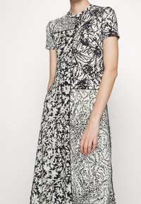 Czarna i biała sukienka w kwiatowy wzór z krótkimi rękawami, okrągłym dekoltem i panelem designem. Materiał wydaje się gładki o lekkiej fakturze.