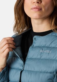 Hellblaue Daunenjacke mit Reißverschluss und gestepptem Design. Mit der aufgestickten Aufschrift 'SEASIDE' auf der Brust. Teils offener Kragen abgebildet.