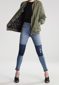 Bomber verde oliva con forro floral, camiseta negra y jeans skinny azules con paneles en bloques de color y detalles desgastados, combinados con tacones negros.