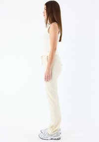 Pantaloni jogger beige con vita elastica e coulisse, abbinati a una canotta aderente color crema. Le scarpe includono delle scarpe da corsa argentate.