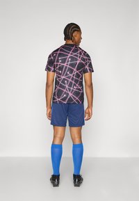 Juoda sportinė marškinė su rožiniu geometriniu raštu, tamsiai mėlynos šortai ir mėlynos kelių ilgio kojinės. Modelis vilki juodus futbolo batelius.