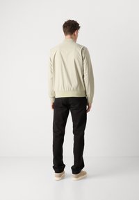 Hellebeige Bomberjacke mit geripptem Saum und Bündchen, kombiniert mit schwarzen Hosen und hellen Sneakern, von hinten betrachtet.