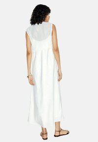 Robe maxi sans manches blanche avec un panneau arrière transparent et des détails en dentelle à la taille. Texture douce, silhouette fluide, assortie à des sandales marron.