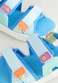 Sandales bleues pour tout-petit avec des brides rayées réglables, des fermoirs carrés colorés et des semelles blanches douces sur un fond clair.