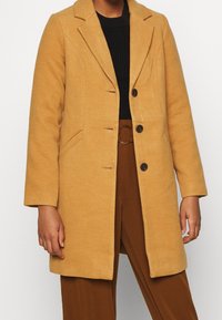 Wool blend coat in mustard yellow with a notched collar, two buttons, and side pockets. Paired with a black sweater and brown trousers.