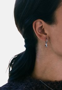 Sidabriniai lankinio auskarai su mažutėle žvaigždės pakabuku. Lankas yra glotnus ir blizgus, o žvaigždės pakabukas turi tekstūruotą paviršių.