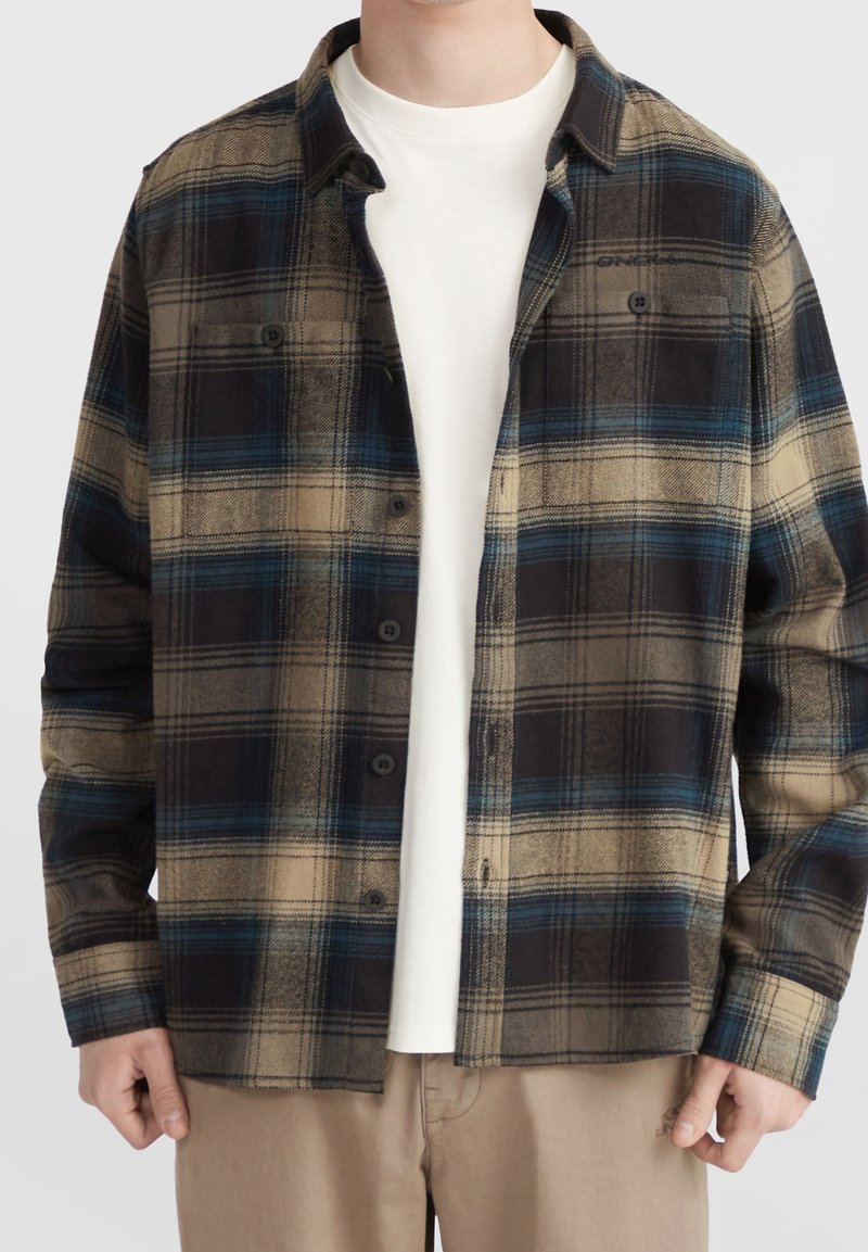 Chemise en flanelle à motifs carreaux marron, noir et bleu. Présente un devant à boutons, deux poches poitrine et des manches longues. Portée par-dessus une chemise blanche.