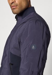 Veste isolante bleu marine avec un col haut, comportant un panneau en maille et un patch logo sur la manche. Texture lisse et design moderne.
