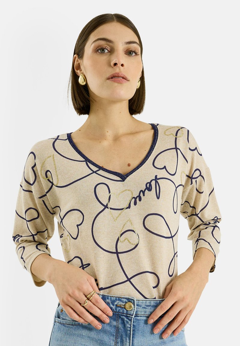 Top beige de manga larga con patrones de corazones en azul marino y dorado, con escote en V y tela texturizada. Combinado con jeans azules de cintura alta.