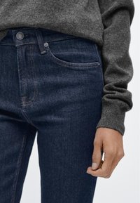 Mörkblå denimjeans med subtilt bleknande, en klassisk design med fem fickor och kontrasterande orangea sömmar. Tyget verkar stadigt och strukturerat.