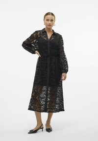 Abito camicia in pizzo nero con motivo floreale, tessuto trasparente, maniche lunghe, chiusura con bottoni, vita stretta e orlo fino al ginocchio.