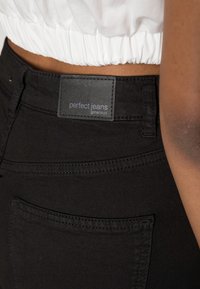 Närbild av en person som bär svarta jeans märkt "perfekta jeans ginatricot" och en vit samlad kort topp.