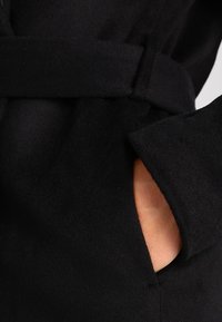 Schwarzer Wollmantel mit einem Taillengürtel, weichem Material und einer seitlichen Tasche mit sichtbarem Handgriff. Der Stoff wirkt glatt und warm.