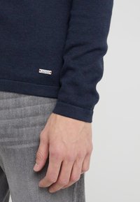 Pull en maille bleu marine avec poignets côtelés et petite étiquette de marque. Porté avec un pantalon gris texturé. Gros plan sur une main posée sur la taille.
