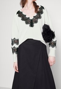 Weißer Pullover mit schwarzem Spitzenbesatz am Halsausschnitt und an den Ärmeln; hat ein strukturiertes schwarzes Fell-Detail. Schwarzer Rock mit Falten.