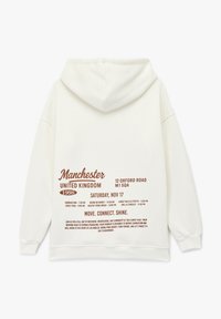 Cremefarbener Hoodie mit einer Kordelzugkapuze. Auf der Rückseite befindet sich in Braun großer gedruckter Text, der eine Veranstaltung in Manchester, Großbritannien, beschreibt.