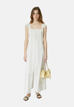 Jeune femme portant une longue robe blanche en dentelle, tenant un petit sac à main beige et portant des sandales noires, debout devant un fond uni.