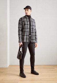 Camicia a quadri in bianco e nero, dolcevita nera, pantaloni neri, scarpe nere. Tiene una piccola borsa nera, in piedi su un pavimento di legno davanti a una parete chiara.