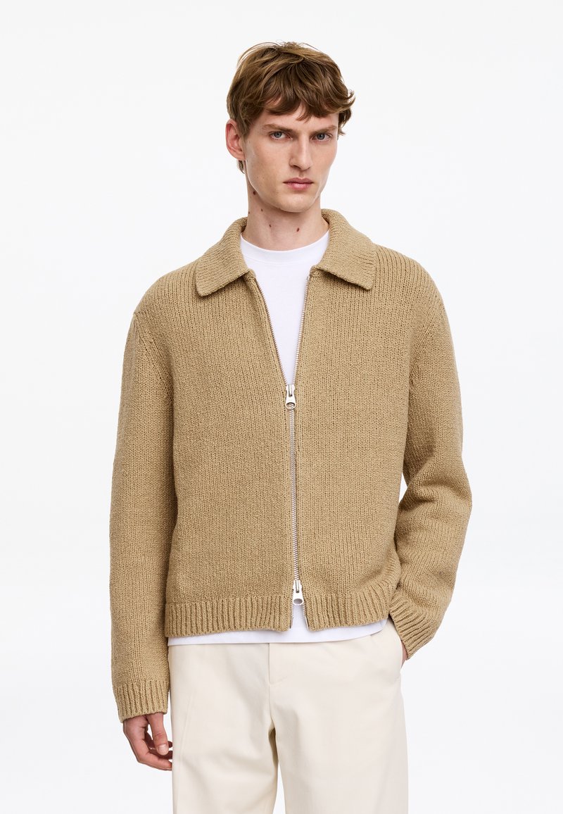 Jeune homme portant un cardigan beige en maille zippé sur une chemise blanche et un pantalon clair, posant avec une main dans la poche.