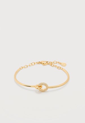 Zlatna narukvica sa malim ukrasnim krugom ukrašenim kristalima i tankim lančanim kopčom na bijeloj pozadini.