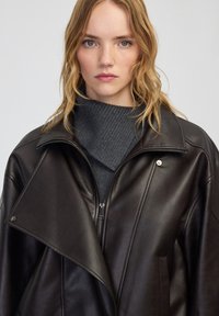 Giacca di pelle marrone scuro con un ampio colletto, chiusura con zip e tasche laterali, indossata sopra un dolcevita a coste grigio scuro.