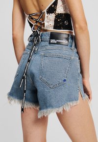 Denim kratke hlače s frayed robom, svetlo modre barve, srednje visoke pasu in črnim blagovno znamko nalepko na hrbtni strani s majhnim modrim logotipom.