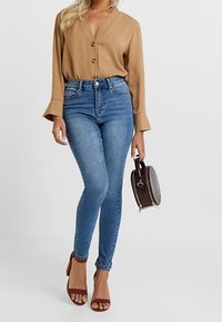Blusa de botón de manga larga color mostaza, jeans ajustados de cintura alta en azul, sandalias de tacón marrones y un pequeño bolso marrón con un diseño texturizado.