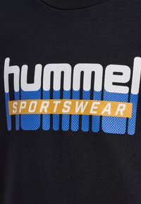 Svart t-shirt med fet "hummel" text i vitt, med blå och gula accenter. Designen inkluderar prickiga blå mönster och en gul banner med "SPORTSWEAR."