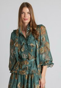 Blusa en verde azulado con bordados florales en dorado y óxido, con volantes, una tapeta de botones y mangas largas con puños fruncidos.
