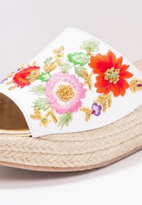 Mule espadrille blanche avec une broderie florale vibrante en rose, rouge et vert. Dispose d'une plateforme en jute tissé et d'une semelle lisse et brillante.