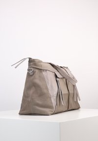 Sac en cuir et suède taupe doux avec des détails de fermeture éclair et de sangle reposant sur une surface blanche contre un fond uni.