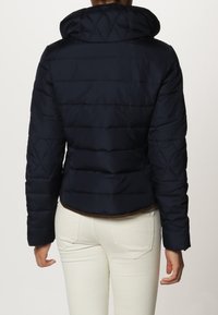 Marineblaue Steppjacke mit Kapuze, aus diamantgefüttertem Material, taillierter Form und elastischem Saum, getragen über hellen Hosen.