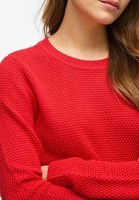 Pull en tricot rouge avec un motif texturé, encolure et poignets côtelés, équipé de manches longues. Gros plan mettant en avant le détail du tissu.