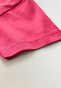 Rosa bomuldsstof med en glat tekstur, med en kant, der er bukket og fladt syet, placeret i en vinkel mod en lys baggrund.