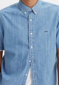 Hombre vistiendo una camisa de mezclilla de manga corta color azul claro con una camiseta blanca debajo, que lleva un pequeño logo de Levi's en el pecho.