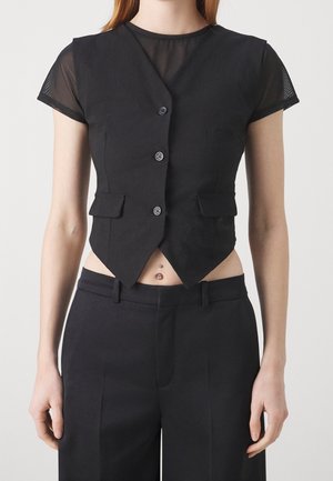 Osoba ubrana w czarną krótko skrojoną kamizelkę z guzikami na przezroczystym czarnym topie z krótkim rękawem i czarnych spodniach, ukazująca kolczyk w pępku.