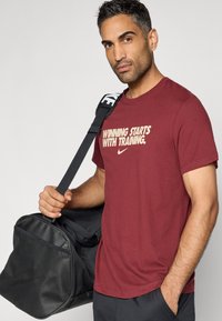 Μπορντό κοντομάνικη μπλούζα με στρογγυλή λαιμόκοψη και το μήνυμα "Η ΝΙΚΗ ΞΕΚΙΝΑ ΜΕ ΤΗΝ ΠΡΟΠΟΝΗΣΗ." τυπωμένο σε μπεζ, συνοδευόμενη από μια μαύρη τσάντα duffel με λουρί.