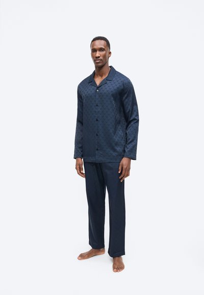 Egy férfi áll mezítláb, sötétkék, hosszú ujjú, gombos felsőt és hozzáillő, laza, mintás nadrágot visel.