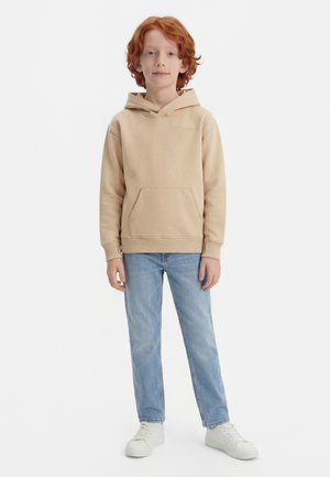 Rødhåret barn iført en beige hættetrøje, lyseblå jeans og hvide sneakers, stående foran en ensfarvet hvid baggrund.