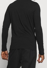 Person, die ein schlichtes schwarzes Langarmshirt und eine schwarze Hose trägt, von hinten gezeigt, mit entspannt an den Seiten hängenden Armen.