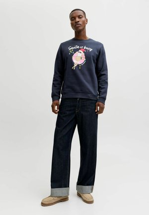 Navy sweatshirt with "Santa is busy" graphic and colorful illustration, paired with loose dark denim jeans and tan shoes. Simple design.