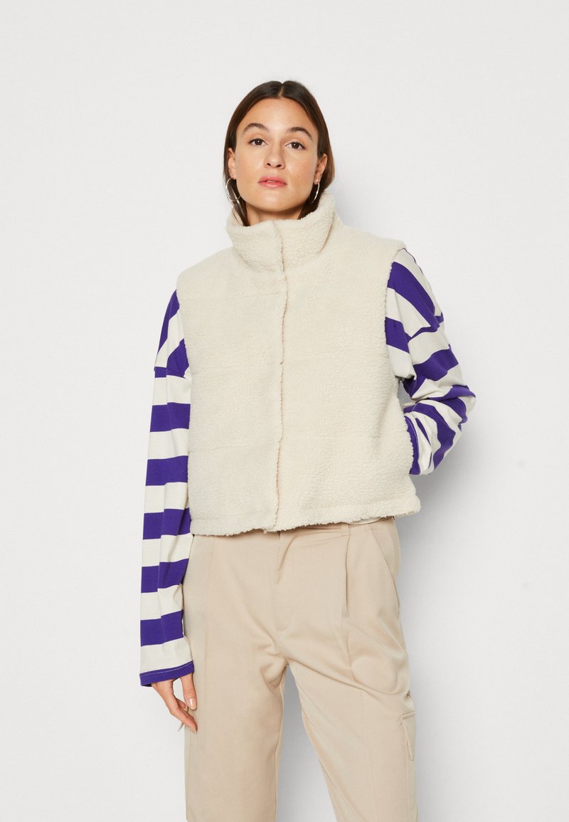 Fleece vest in roomkleur met een hoge kraag en rits aan de voorkant. Gedragen over een paars en witte gestreepte blouse met lange mouwen, gecombineerd met beige broek.