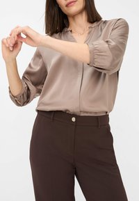 Satin blouse in a muted taupe color, featuring a V-neck and gathered sleeves. Paired with brown trousers, showcasing a smooth texture and fitted design.