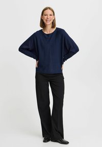 Pull ample bleu marine à manches longues et col rond, associé à un pantalon large noir et des chaussures noires élégantes.