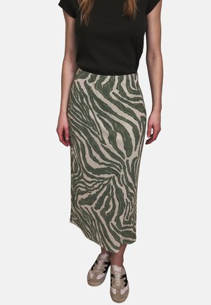 Frau trägt ein schwarzes Kurzarmoberteil, einen knielangen Rock mit grünen und beigen abstrakten Zebrastripes und beigefarbene Turnschuhe mit schwarzen Akzenten.