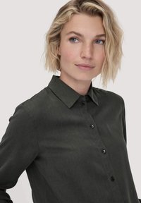 Camicia a maniche lunghe di un verde scuro, con colletto, chiusura frontale con bottoni e un tessuto leggermente testurizzato. Presenta bottoni neri lungo la pattina.