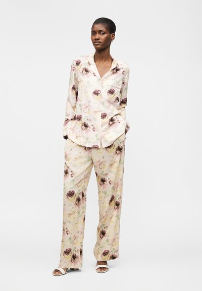 Γυναίκα φοράει ένα φαρδύ, κρεμ χρωματιστό σετ πυτζάμες με μακρύ μανίκι και παντελόνια με φαρδύ πόδι, συνδυασμένο με λευκά σανδάλια ανοιχτής μύτης.