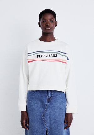 Sudadera corta blanca con el texto "PEPE JEANS" y rayas horizontales multicolor. Lleva puesta una falda de mezclilla azul con una abertura.